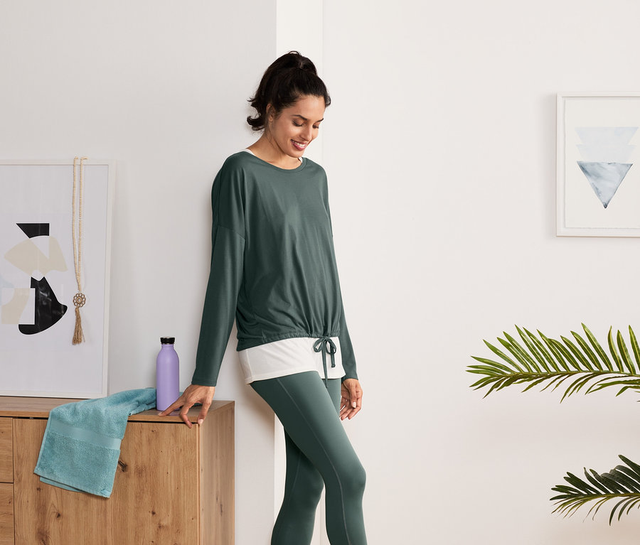 Frau steht an Kommode gelehnt und trägt ein grün-weißes 2-in-1-Sportshirt.