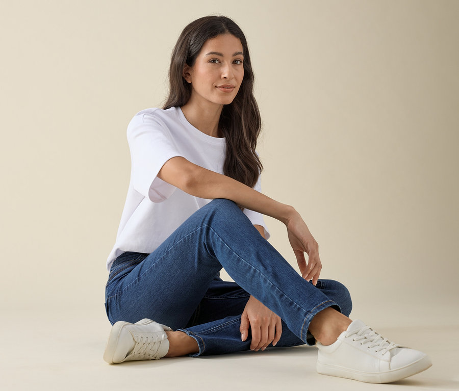 Frau sitzt am Boden, trägt Slim-Jeans – Fit »Emma« in Mid Blue Denim, ein weißes T-Shirt und Sneakers.