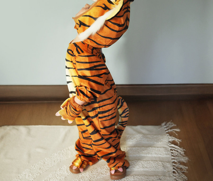 Ein Kind im Small Foot Kostüm „Tiger“ steht auf einem Teppich.