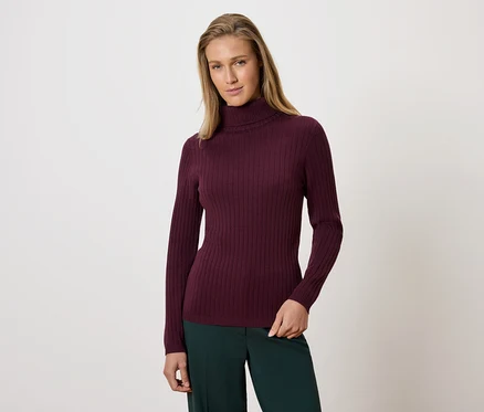 Frau posiert in einem bordeauxroten Rollkragenpullover und einer grünen Hose.