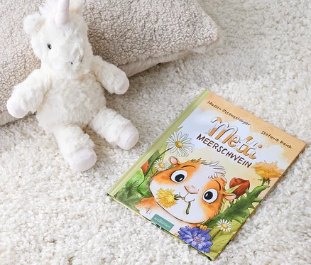 Ein Plüsch-Einhorn liegt neben dem Buch »Metti Meerschwein« auf einem weißen Teppich.