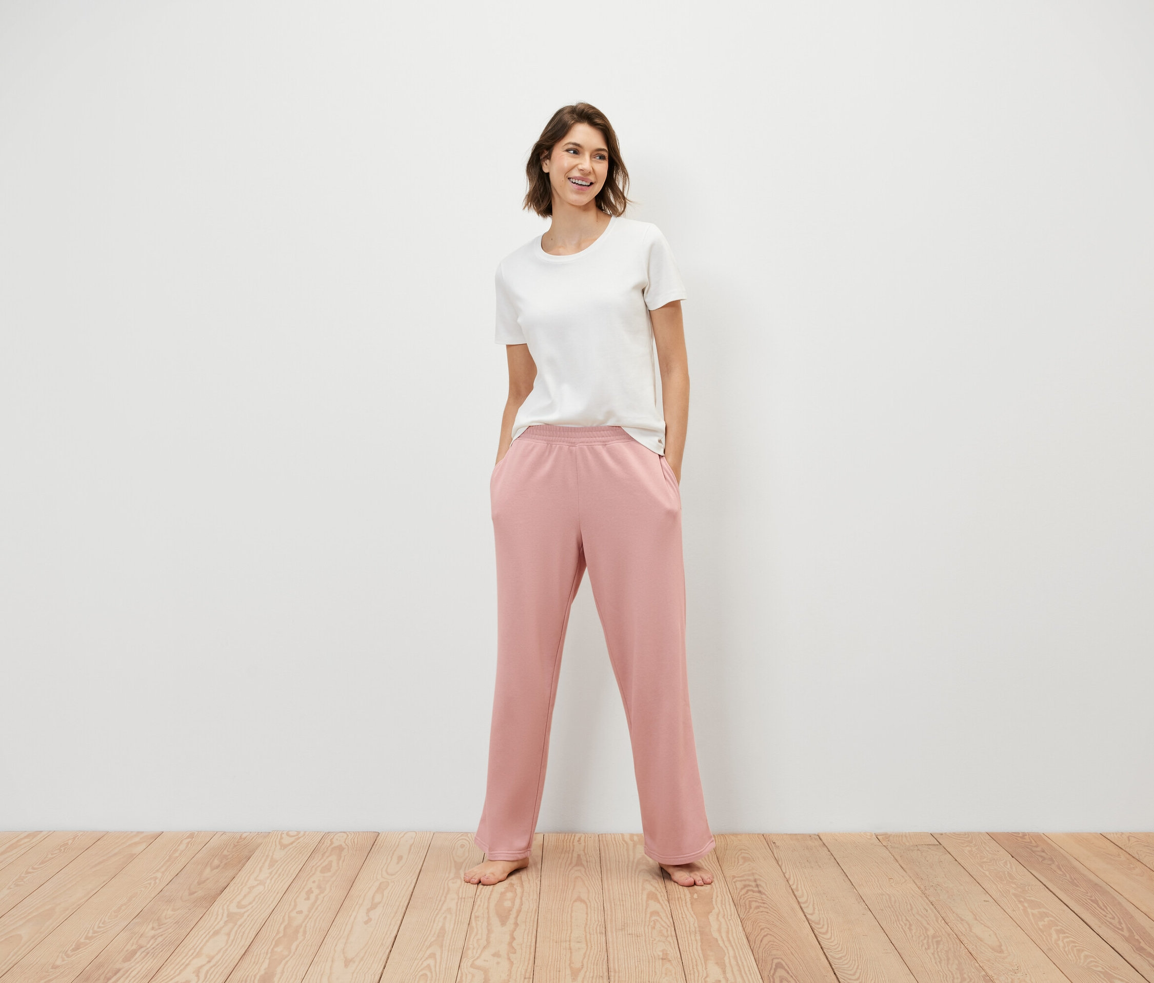 Frau steht mit Händen in den Taschen einer rosafarbenen Jogginghose und trägt ein weißes T-Shirt.