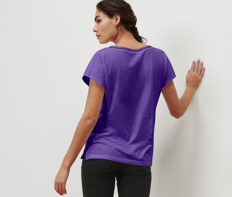 Frau mit geflochtenen Haaren, von hinten, an eine Wand gelehnt, trägt ein lila Shirt aus Modalmix.