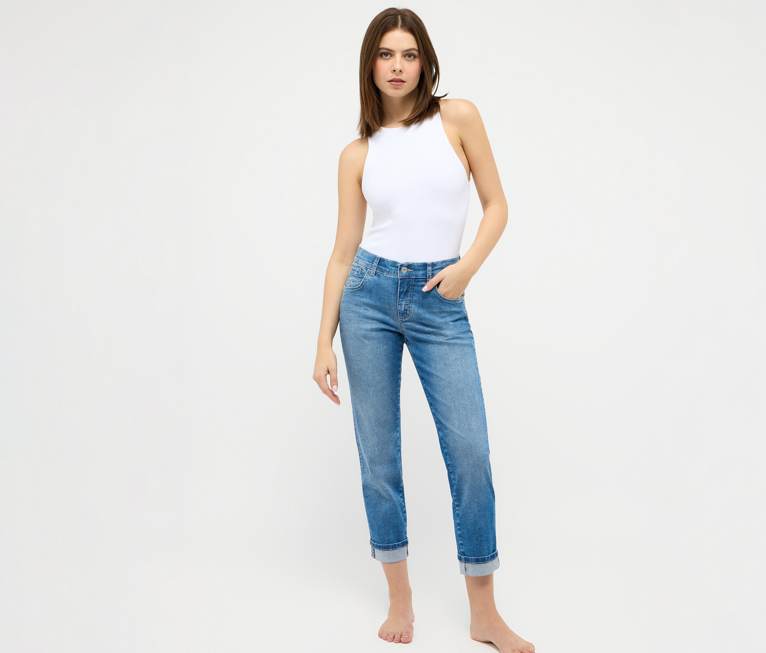 Frau steht barfuß vor einem weißen Hintergrund und trägt eine weiße Bluse und eine Jeans »Darleen« Crop Turn Up 7/8 von Angels in Hellblau.