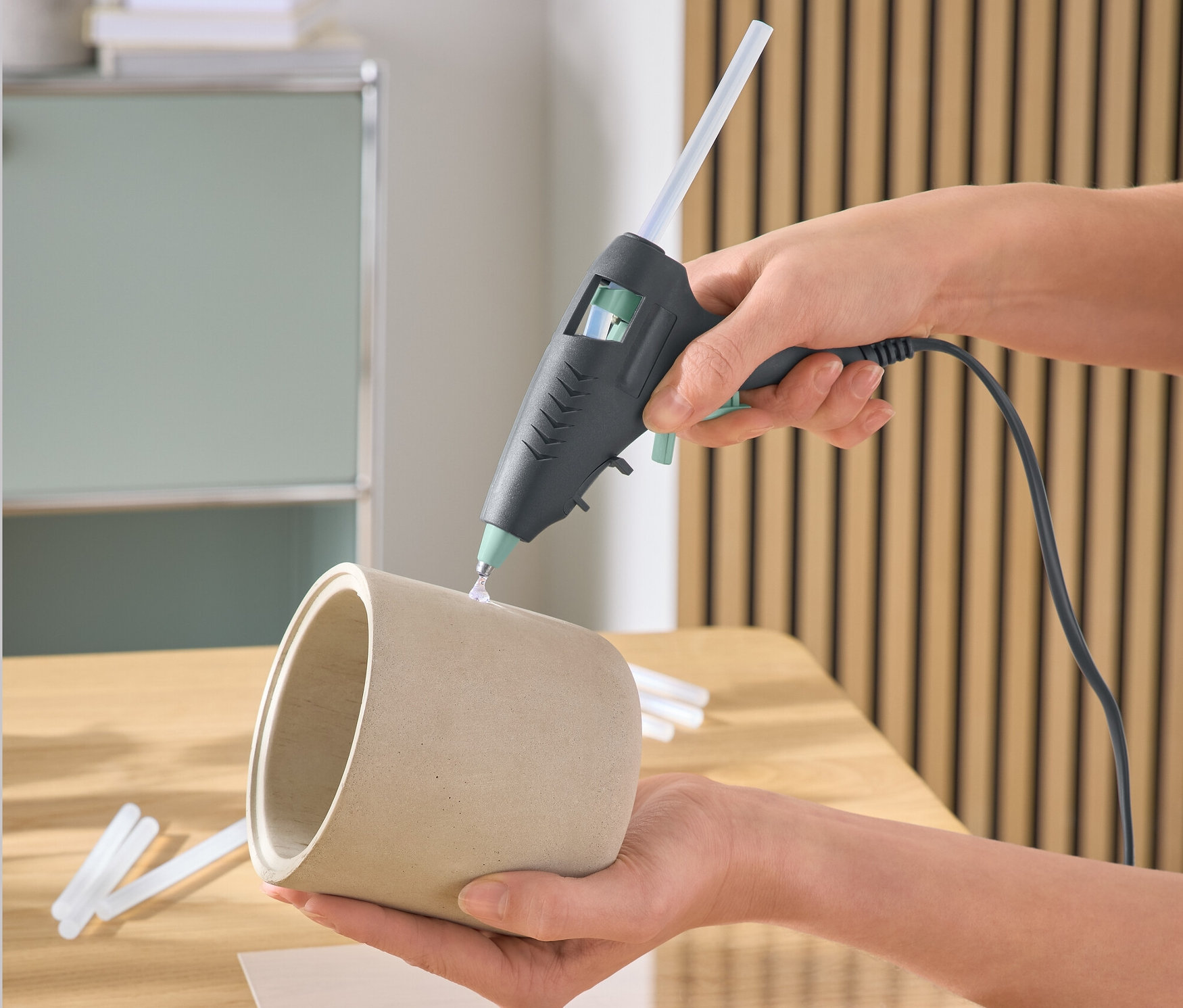 Eine Hand benutzt eine Klebepistole, um einen Blumentopf zu bekleben. Auf dem Tisch liegen Muscheln.