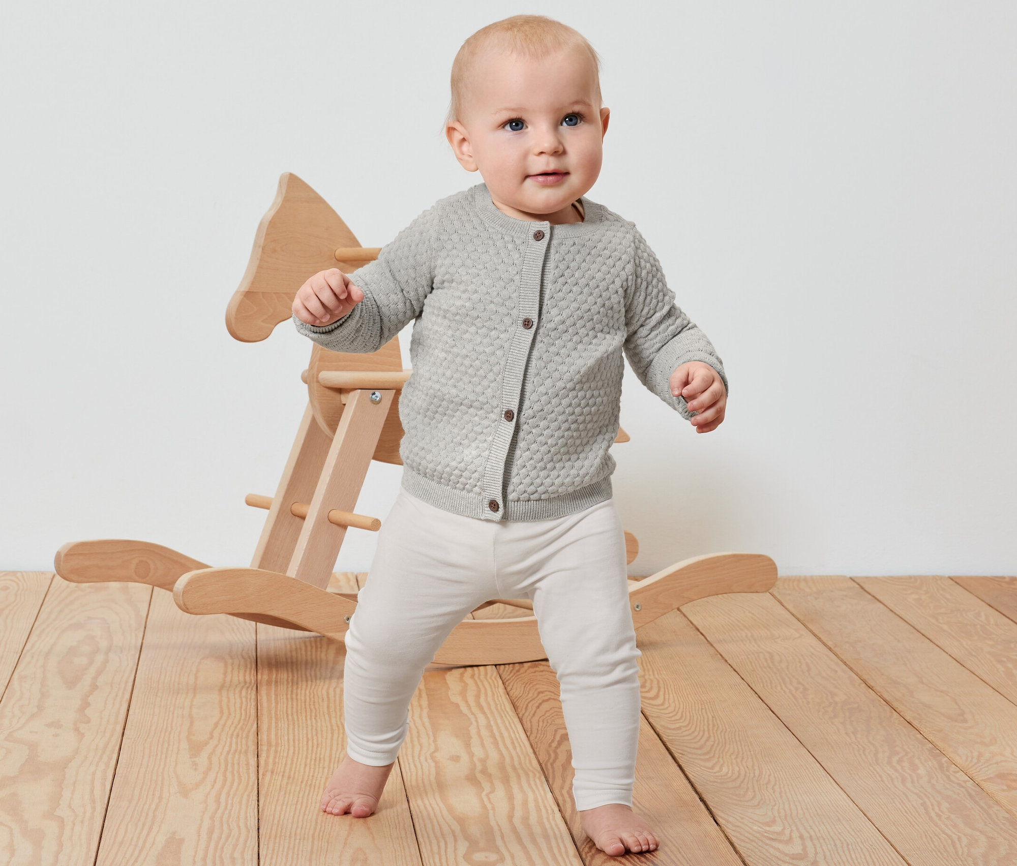 Baby steht neben einem Schaukelpferd aus Holz und trägt eine Baby-Strickjacke.