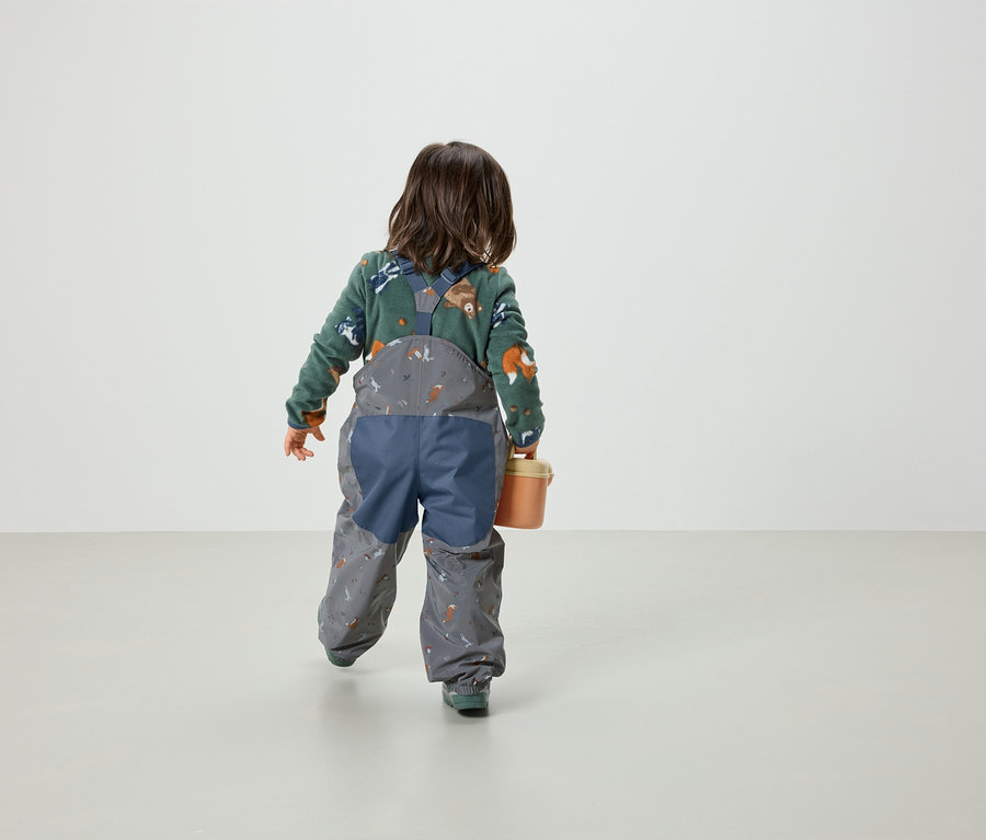 Kleinkind von hinten in einem grünen Fuchspullover und grauen Latzhosen mit Print hält einen orangefarbenen Eimer.