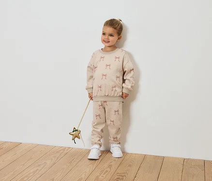 Ein lächelndes kleines Mädchen hält einen sternförmigen Zauberstab und trägt ein beiges Outfit mit Schleifenmuster.