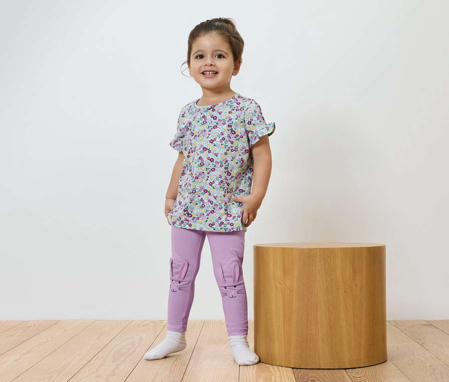 Ein lächelndes kleines Mädchen steht vor einem braunen zylinderförmigen Tisch und trägt ein geblümtes T-Shirt und lila Leggings.