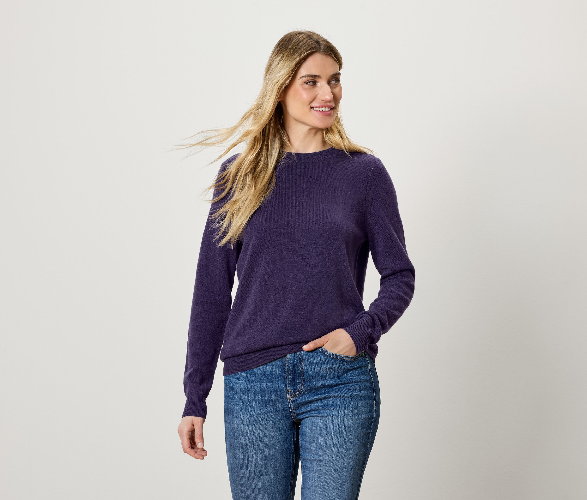 Frau mit blonden Haaren trägt einen violetten Pullover und blaue Jeans.