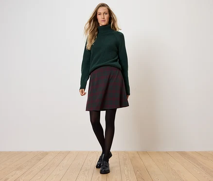 Frau steht und trägt einen grünen Rollkragenpullover, einen karierten Rock, eine schwarze Strumpfhose und schwarze Schuhe.
