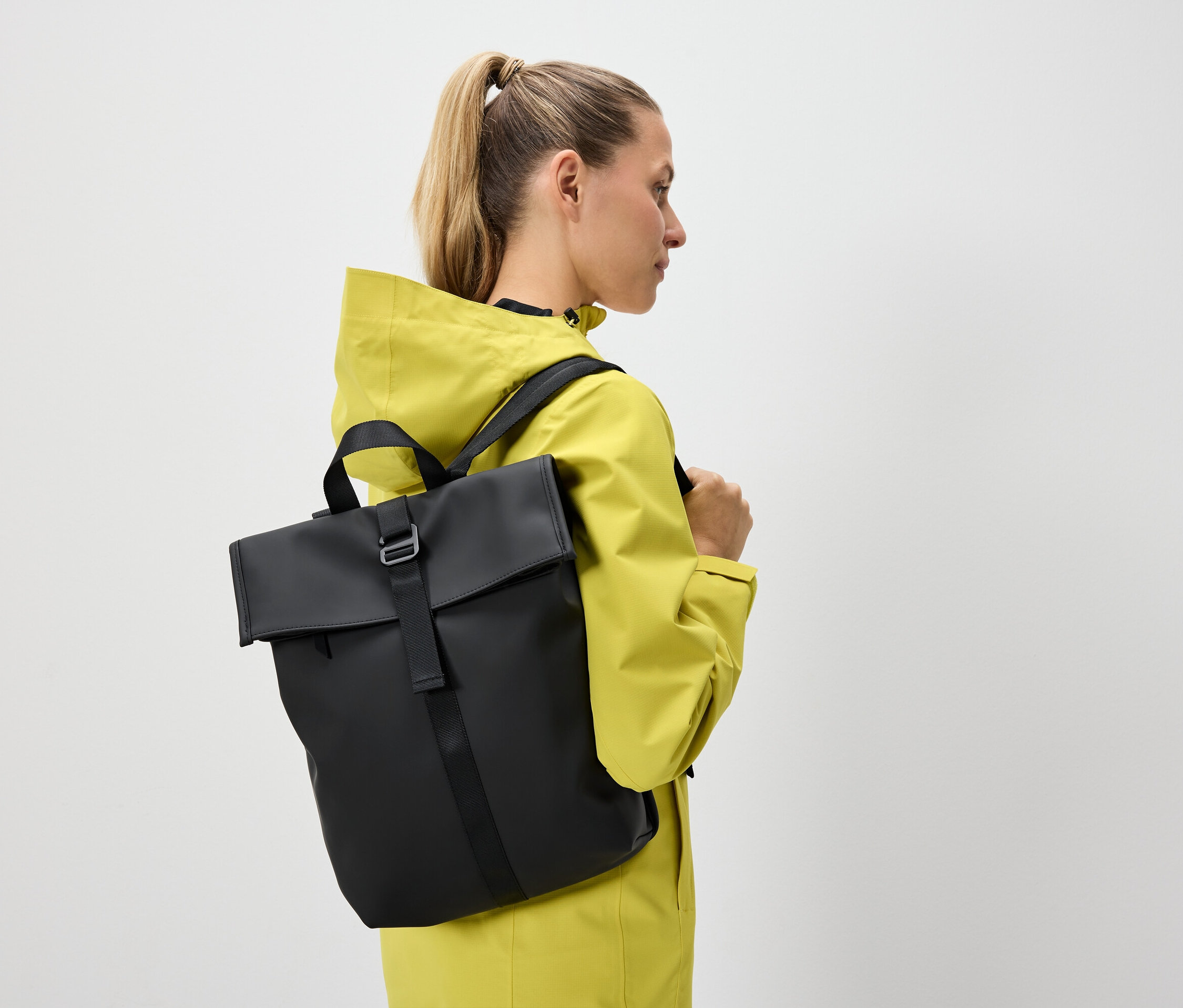 Frau mit blonden, zum Pferdeschwanz gebundenen Haaren trägt eine gelbe Jacke und einen schwarzen Rucksack.