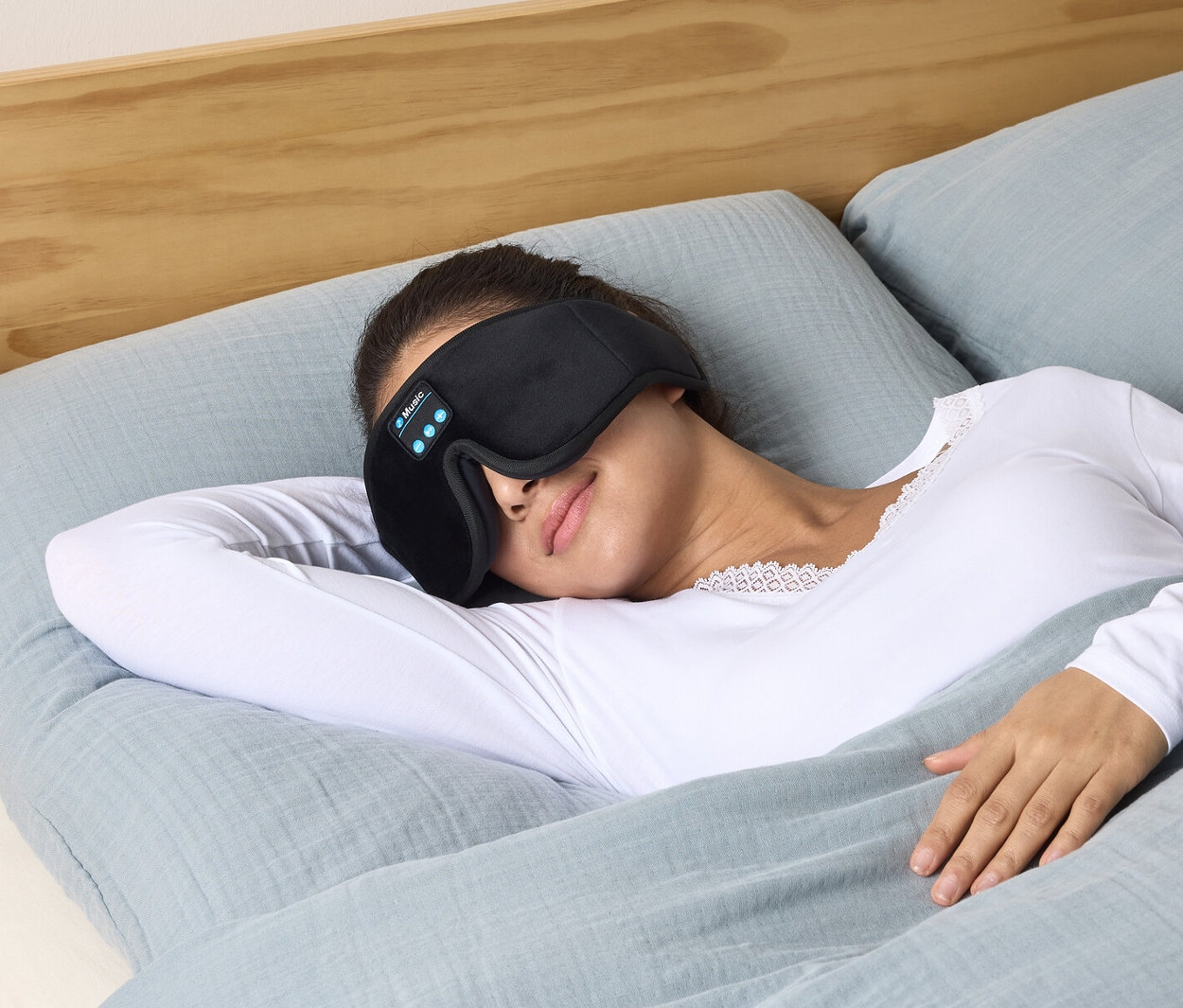 Frau liegt im Bett und trägt eine Schlafmaske mit Kopfhörern.