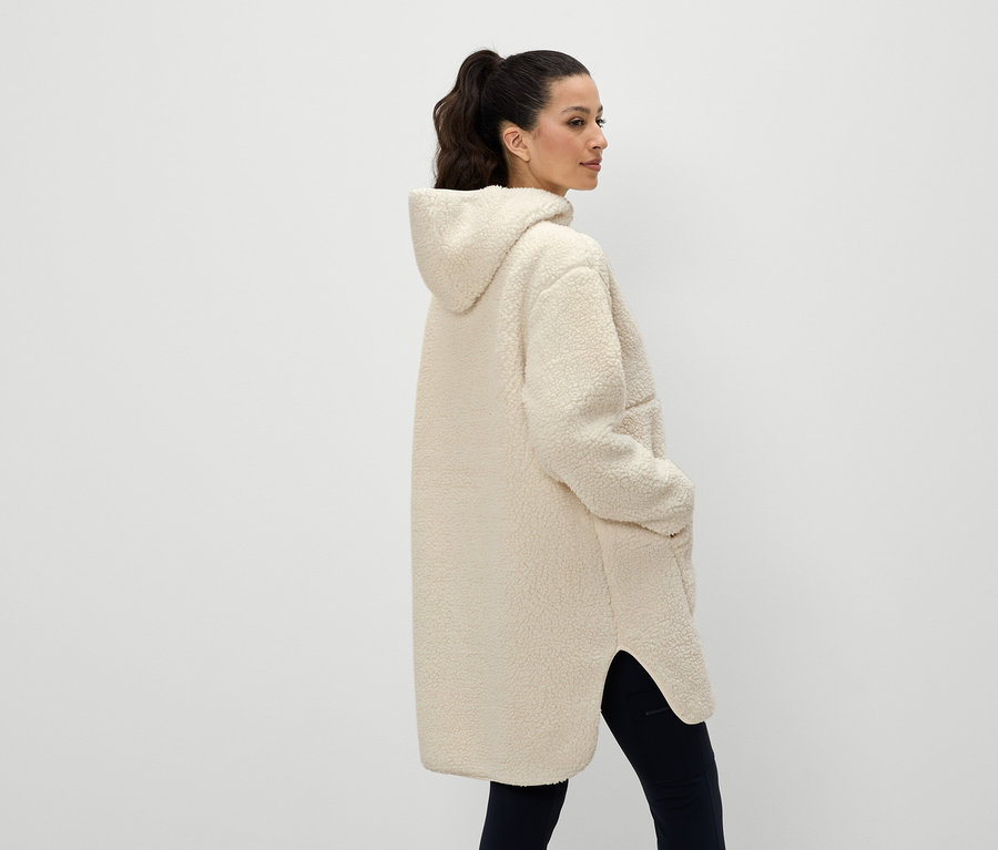 Eine Frau von hinten trägt einen cremefarbenen Kapuzenmantel und hat die Hände in den Taschen.