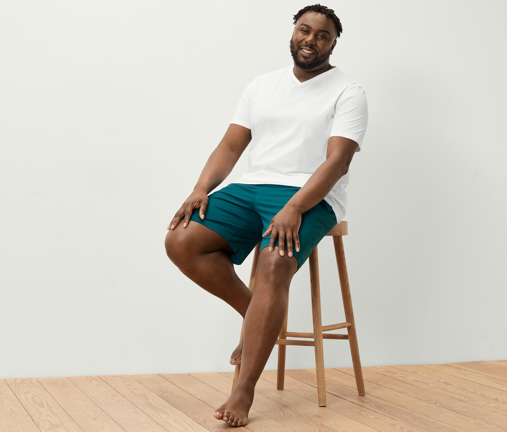 Ein Mann sitzt auf einem Hocker und trägt ein weißes T-Shirt mit V-Ausschnitt und Relaxshorts.