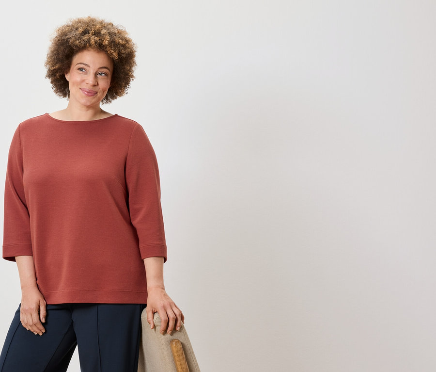 Frau mit lockigem Haar posiert in einem rotbraunen Pullover und dunkelblauen Hosen.