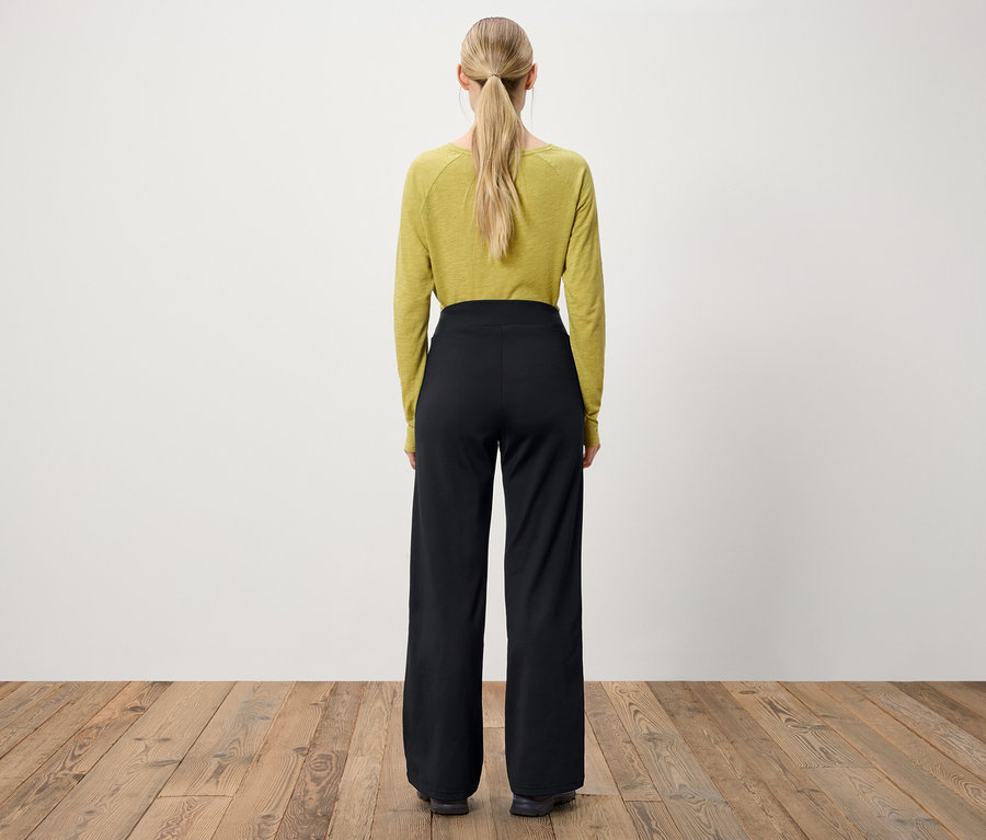 Von hinten ist eine Frau mit blonden Haaren zu sehen, die zu einem Pferdeschwanz zusammengebunden sind. Sie trägt ein olivgrünes Langarmshirt und eine schwarze Hose mit weitem Bein.
