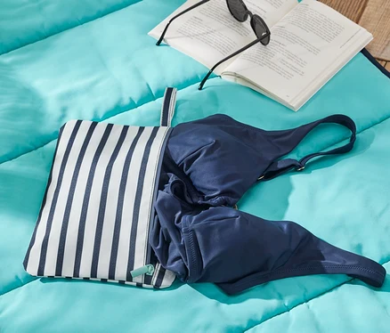 Auf einer blauen Decke liegen ein Nassbeutel, eine blaue Badehose, ein Buch und eine Sonnenbrille.