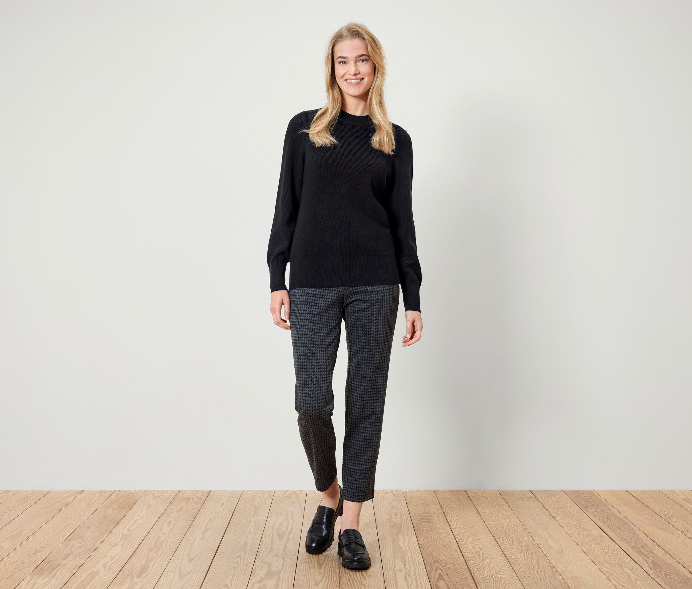 Eine Frau steht und lächelt, sie trägt einen schwarzen Pullover, gemusterte Hosen und schwarze Slipper.