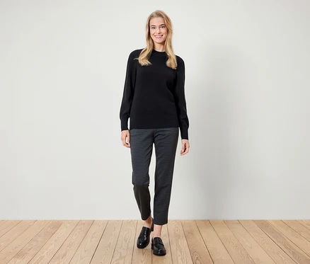 Eine Frau steht und lächelt, sie trägt einen schwarzen Pullover, gemusterte Hosen und schwarze Slipper.