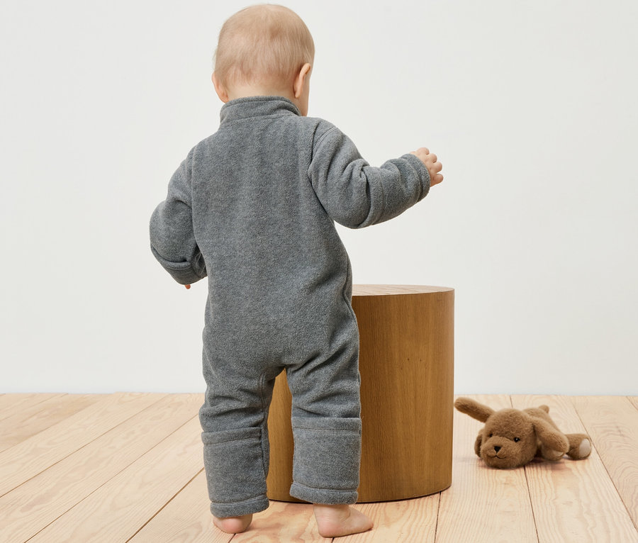 Ein Baby in einem grauen Fleece-Einteiler steht neben einem Holzhocker. Ein Stoffhund liegt daneben.