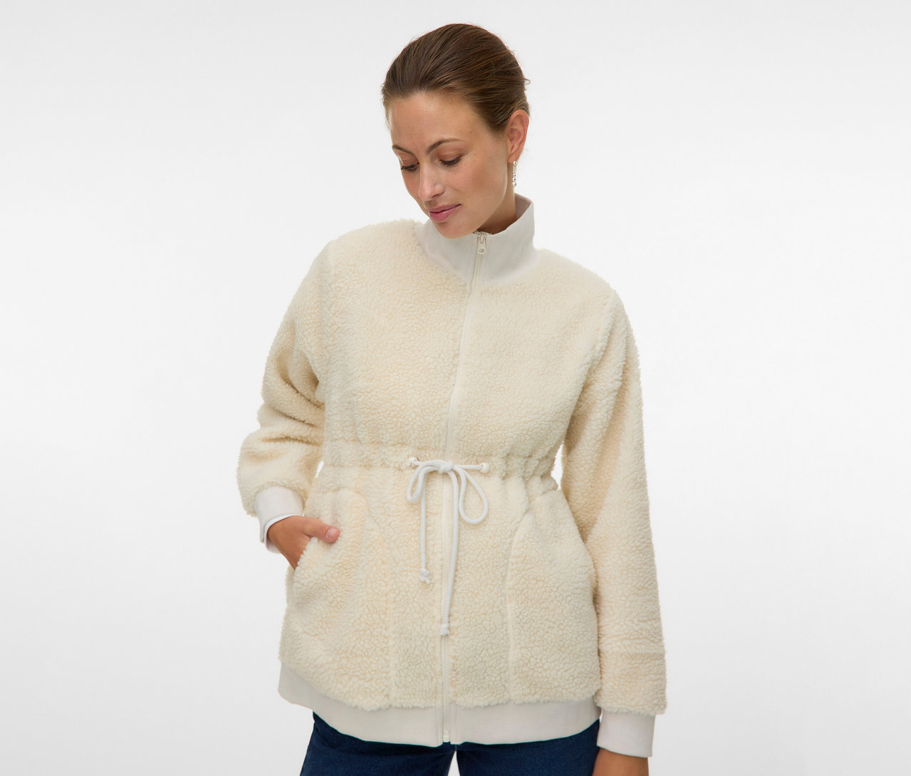 Frau mit der offenen, cremefarbenen MAMALICIOUS 3-in-1-Teddyjacke »MLCARLY«, Kordelzug in der Taille und Hände in den Taschen.