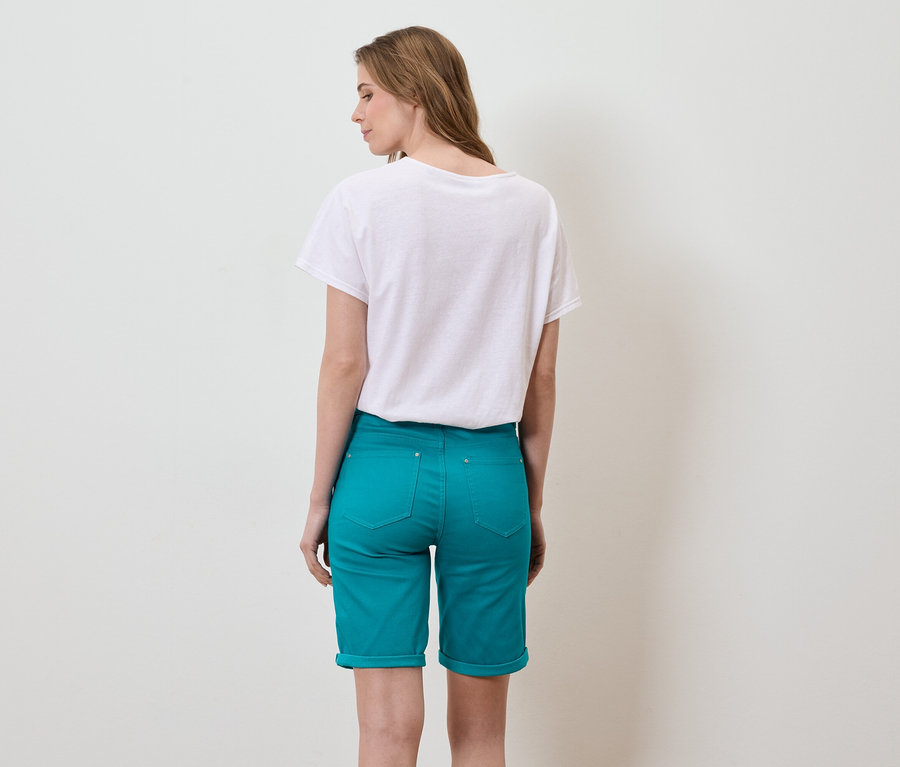 Eine Frau mit langen braunen Haaren steht mit dem Rücken zur Kamera und trägt ein weißes T-Shirt und türkisfarbene Shorts.