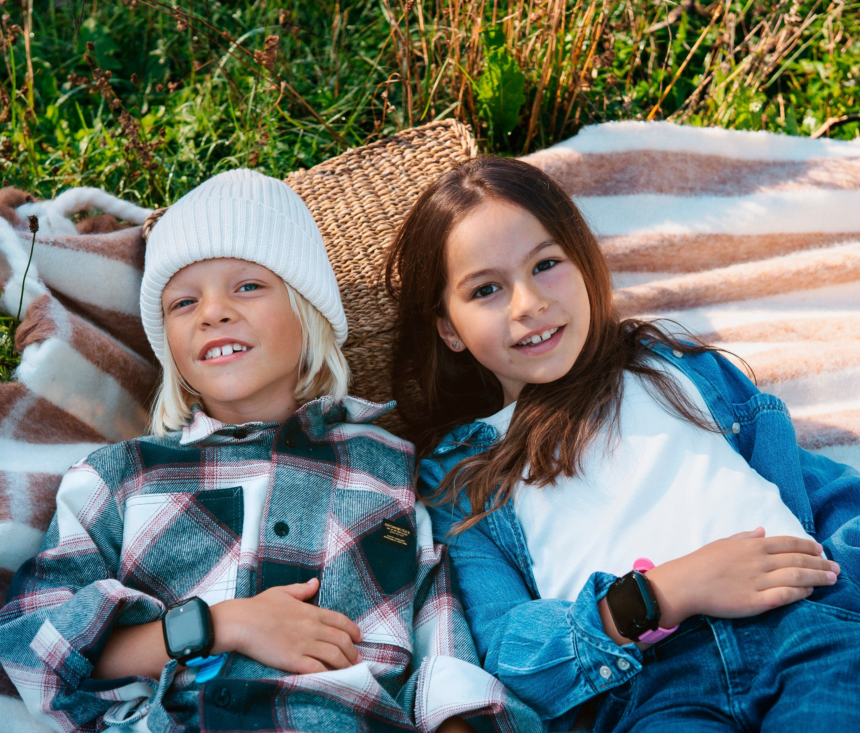 Zwei Kinder liegen auf einer gestreiften Decke im Gras. Ein Kind trägt eine pinke Kids Watch Xplora XGO2.