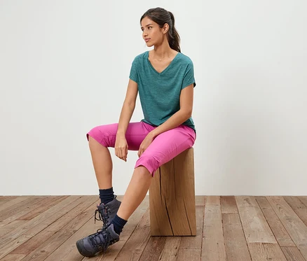 Frau sitzt auf einem Holzschemel, trägt ein grünes T-Shirt, pinke Caprihosen und Wanderschuhe.