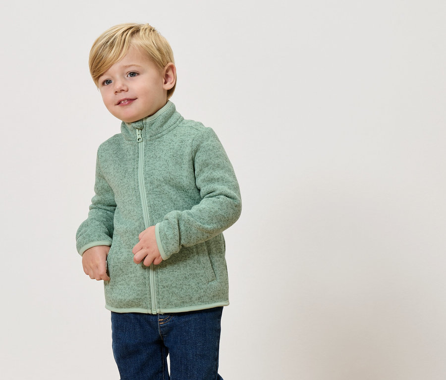 Ein blonder Junge trägt eine hellgrüne Fleecejacke und Jeans.