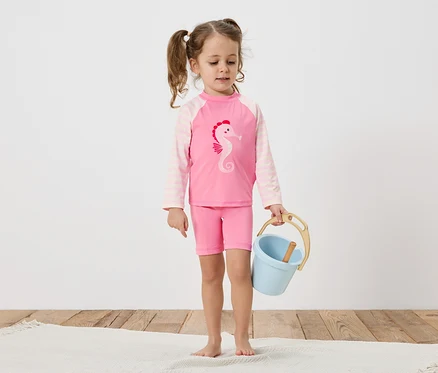 Ein kleines Mädchen mit Zöpfen trägt einen rosa Badeanzug mit Seepferdchen-Motiv und hält einen blauen Eimer auf einem Holzboden.