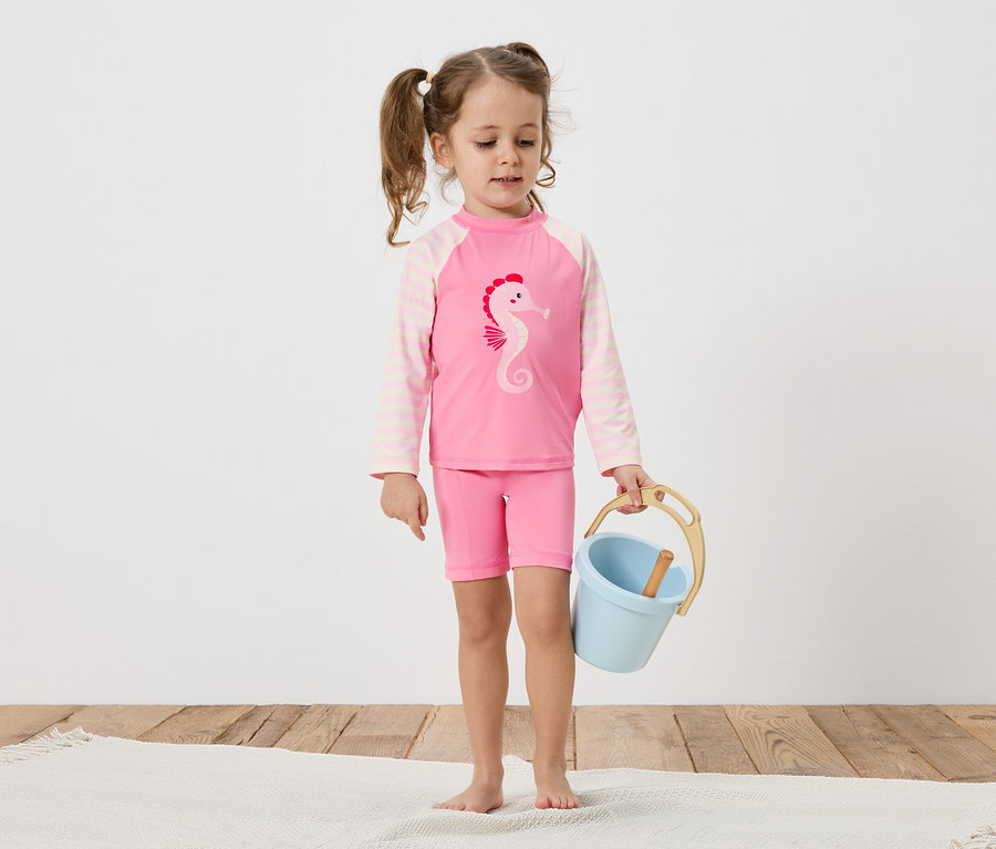 Ein kleines Mädchen mit Zöpfen trägt einen rosa Badeanzug mit Seepferdchen-Motiv und hält einen blauen Eimer auf einem Holzboden.