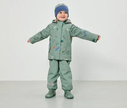 Ein Kleinkind in hellgrüner Regenbekleidung mit Dinosaurier-Muster und blauer Mütze steht mit ausgestreckten Armen vor einem weißen Hintergrund.