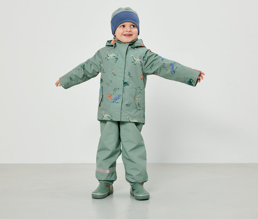 Ein Kleinkind in hellgrüner Regenbekleidung mit Dinosaurier-Muster und blauer Mütze steht mit ausgestreckten Armen vor einem weißen Hintergrund.