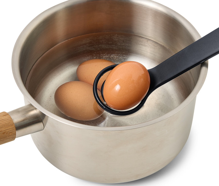 Ein braunes Ei wird mit einem schwarzen Eierlöffel aus einem Topf mit drei Eiern gehoben.