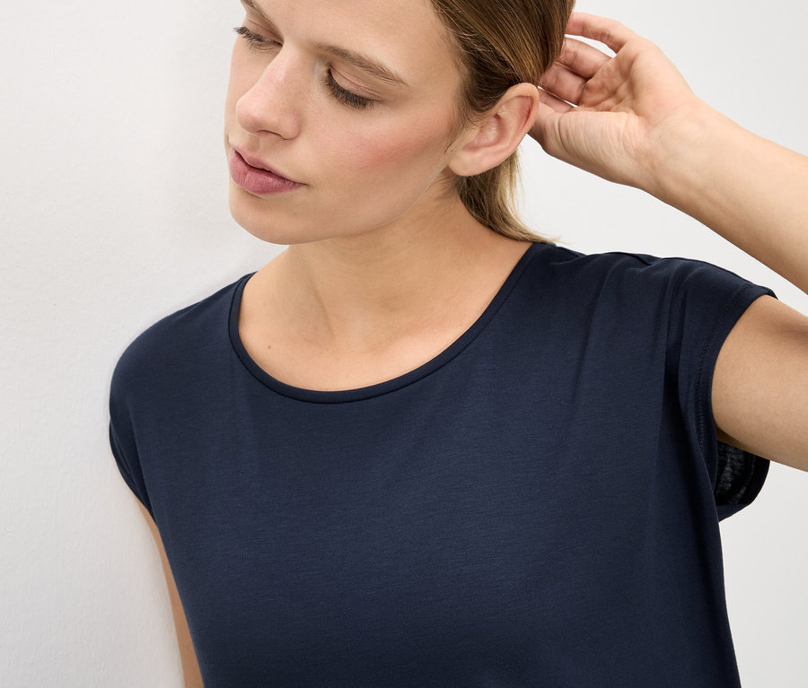 Frau mit dunklem T-Shirt berührt ihr Ohr.