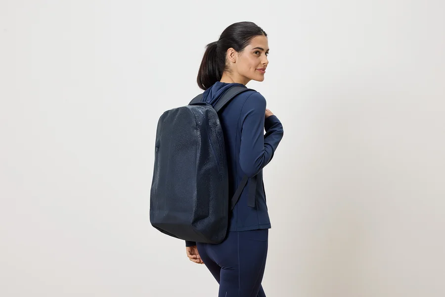 Frau mit marineblauem Trainingsanzug und marineblauem Rucksack, der über ihre Schulter hängt, blickt über ihre Schulter.