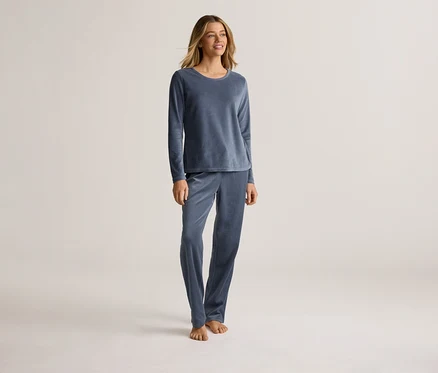 Eine Frau steht barfuß in einem blauen, langärmeligen Samtpyjama vor einem hellen Hintergrund.