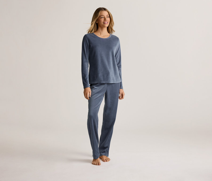 Eine Frau steht barfuß in einem blauen, langärmeligen Samtpyjama vor einem hellen Hintergrund.