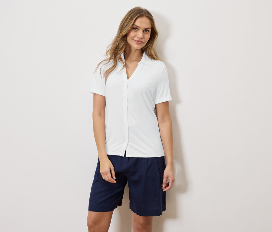 Eine Frau steht vor einer Wand und trägt ein weißes Hemd und blaue Shorts.