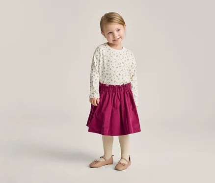 Ein kleines Mädchen mit blonden Haaren trägt ein langärmliges Oberteil mit Blumenmuster, einen roten Rock, Strumpfhosen und Ballerinas.