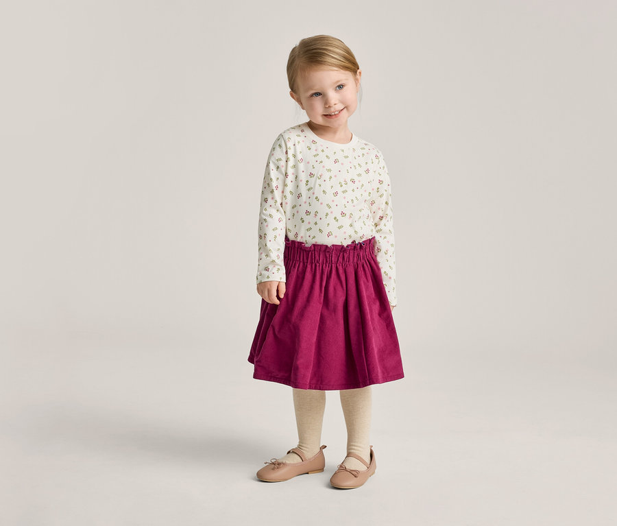 Ein kleines Mädchen mit blonden Haaren trägt ein langärmliges Oberteil mit Blumenmuster, einen roten Rock, Strumpfhosen und Ballerinas.