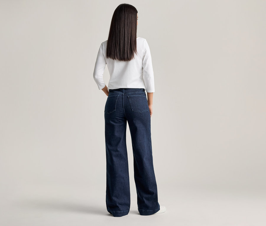Rückansicht einer Frau mit langen, dunklen Haaren, die eine weiße Bluse und eine weite, dunkelblaue Jeans trägt.