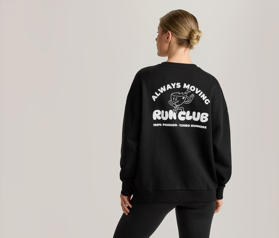 Eine Frau von hinten trägt ein schwarzes Sweatshirt mit einem weißen Aufdruck. Der Aufdruck zeigt einen Cartoon-Kaffeebecher und den Text 'ALWAYS MOVING RUN CLUB' sowie '100% PASSION - TCHIBO RUNNERS'.