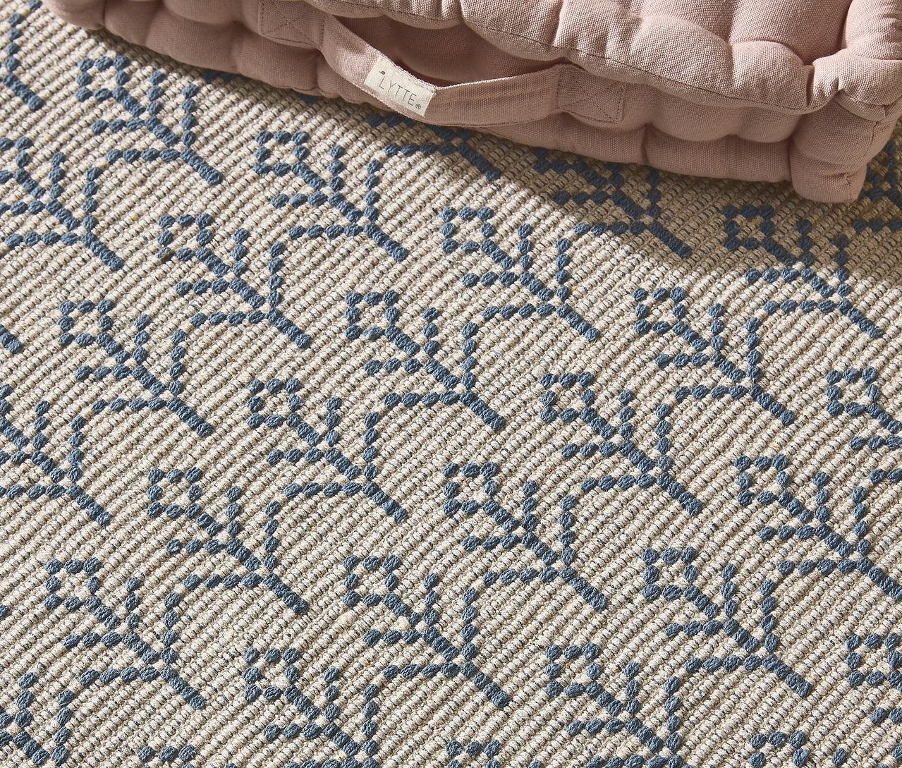 Nahaufnahme eines beigen Teppichs mit blauem Muster und einem rosa Kissen in der Ecke.