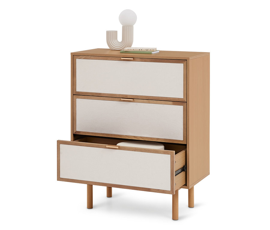 Helle Holzkommode mit drei cremefarbenen Stoffschubladen und goldenen Griffen. Die unterste Schublade ist geöffnet und zeigt ein weißes Objekt. Eine Dekoleuchte und Bücher stehen oben.
