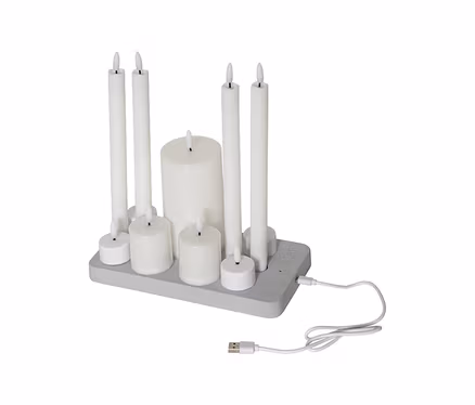 Neun weiße LED-Kerzen unterschiedlicher Größen, darunter Stumpen- und Teelichtkerzen, auf einer grauen Ladestation mit angeschlossenem USB-Kabel.