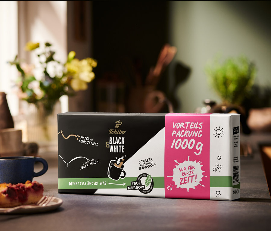 Eine Packung BLACK & WHITE - 1 kg Gemahlen Kaffee steht neben einem Stück Kuchen und einer blauen Tasse.