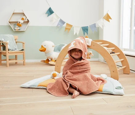 Lächelndes Kind, eingewickelt in ein pfirsichfarbenes Kapuzenhandtuch mit Entenmotiv, sitzt barfuß auf einer Regenbogen-Spielmatte in einem Zimmer mit Schaukelente und Kletterbogen.