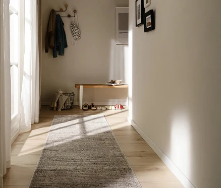 Heller Flur mit benuta Läufer »Anwar« in Grau-Blau auf Holzboden. Sonnenlicht fällt durch ein Fenster. Eine Garderobe mit Jacken hängt über einer Holzbank mit Schuhen und Korb.